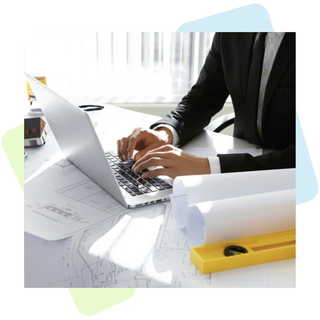 A businessman in a black suit typing on a laptop at a desk covered in construction plans and rolled blueprints, illustrating Why Choose Remire to Provide Contractor Management Services to Your Company.