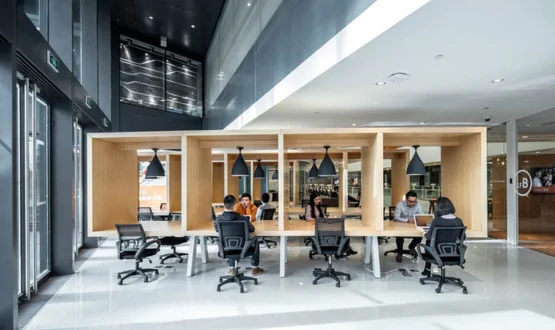 A modern, open-concept corporate office interior showcasing ScaleBridge improved contractor management strategies by Remire. The image depicts several professionals working with laptops at a long shared wooden desk equipped with integrated box-shaped cubicle dividers and low-hanging black pendant lights in a bright, high-ceilinged coworking space.